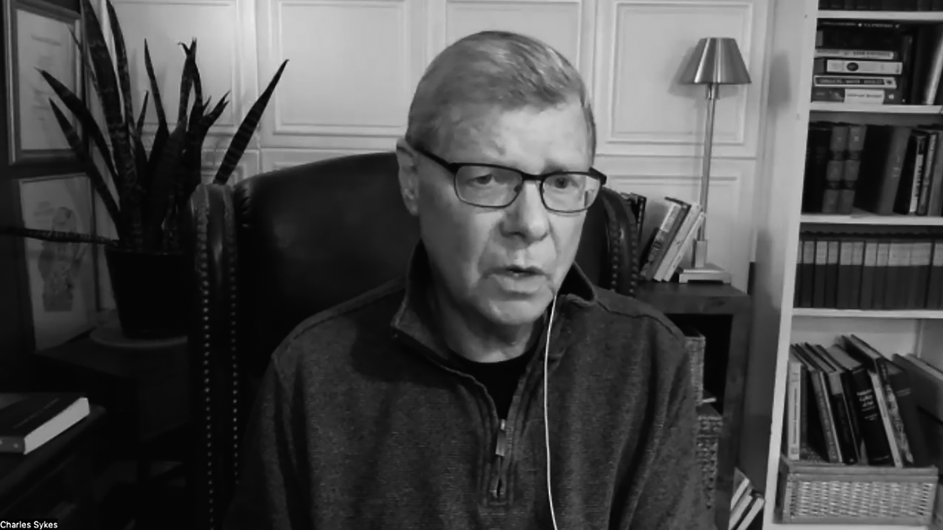 A grayscale screenshot with the text "Charlie Sykes" in the bottom left corner shows Charlie Sykes with a corded headphone in his left ear while sitting in a high-backed leather chair in a room with a bookcase, table with a lamp, potted plant and framed items on a wall.