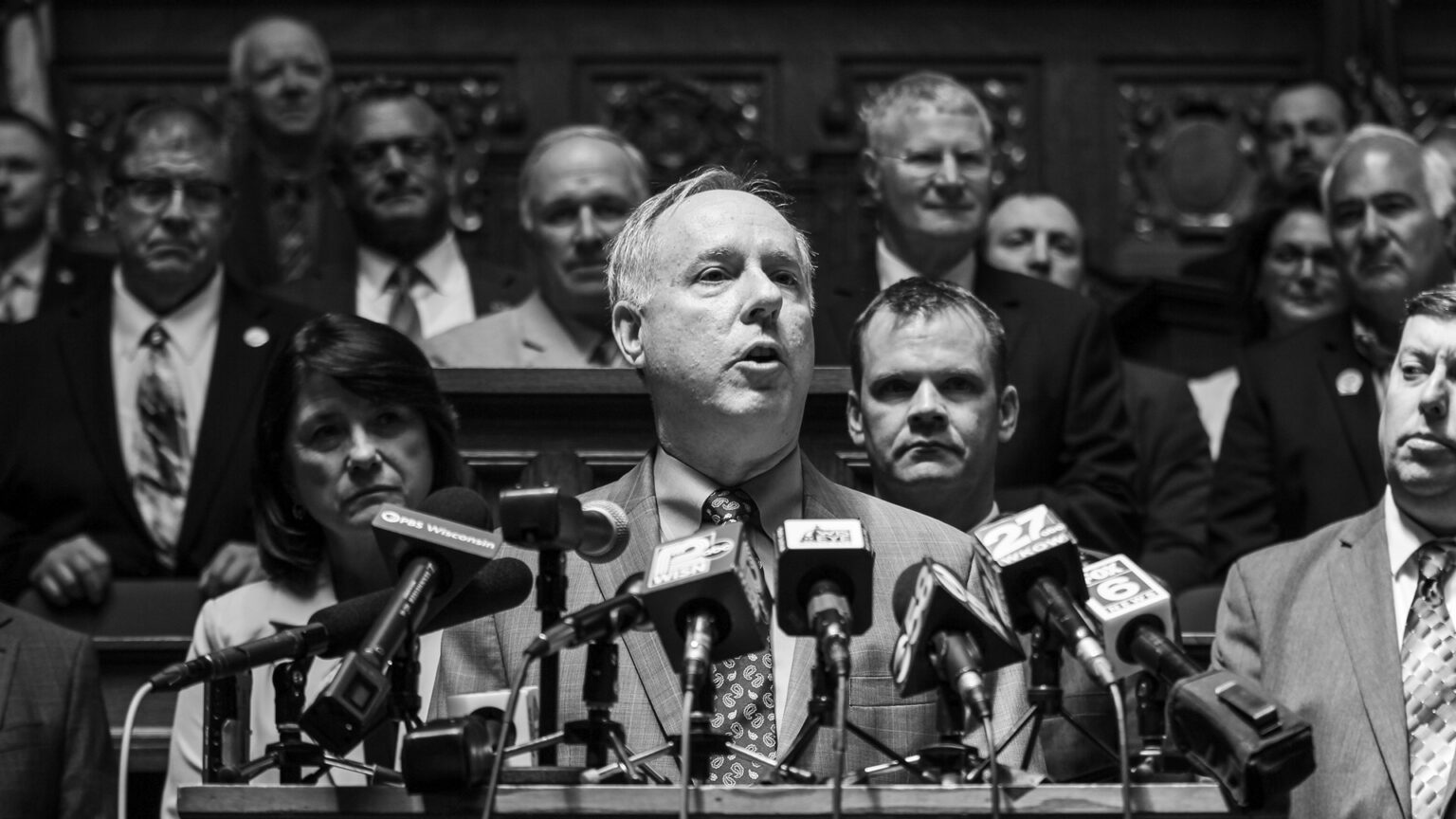 A grayscale photo shows Robin Vos standing and speaking into nine microphones with the flags of different media organizations mounted to the top of a wood podium, with other people standing behind him on multiple levels of a legislative dais.