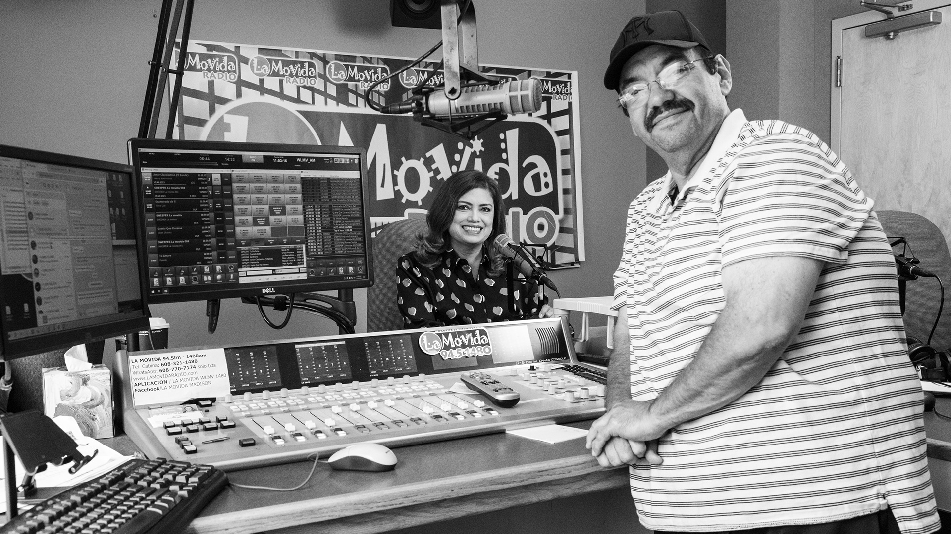 Lupita Montoto poses for a portrait while sitting in a chair and faces a mounted microphone in front of a poster for "La Movida" and behind a desk with an audio mixing board, multiple monitors and keyboard on its surface, with Luis Montoto posing for a portrait while standing in front of the table and facing a microphone on a boom stand.