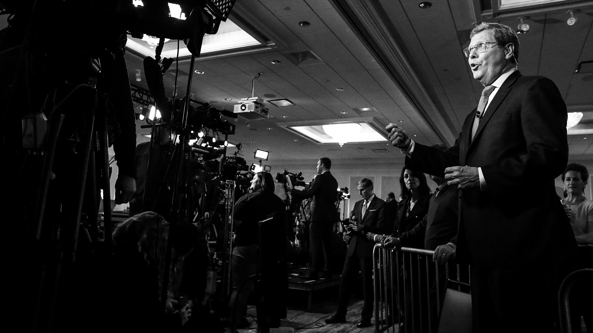 A grayscale photo shows Charlie Sykes standing and speaking while gesturing with his right hand and facing multiple television cameras, with other people and cameras in the background in a room with recessed chandeliers in a drop ceiling.