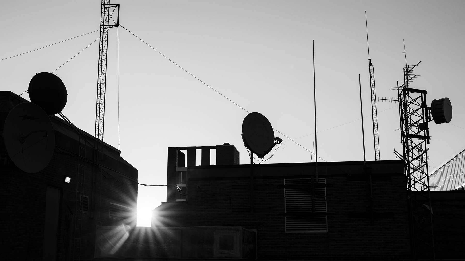 A grayscale photo shows satellite dishes and antennas on top of a roof, with sunshine rays visible between two raised portions of the building.