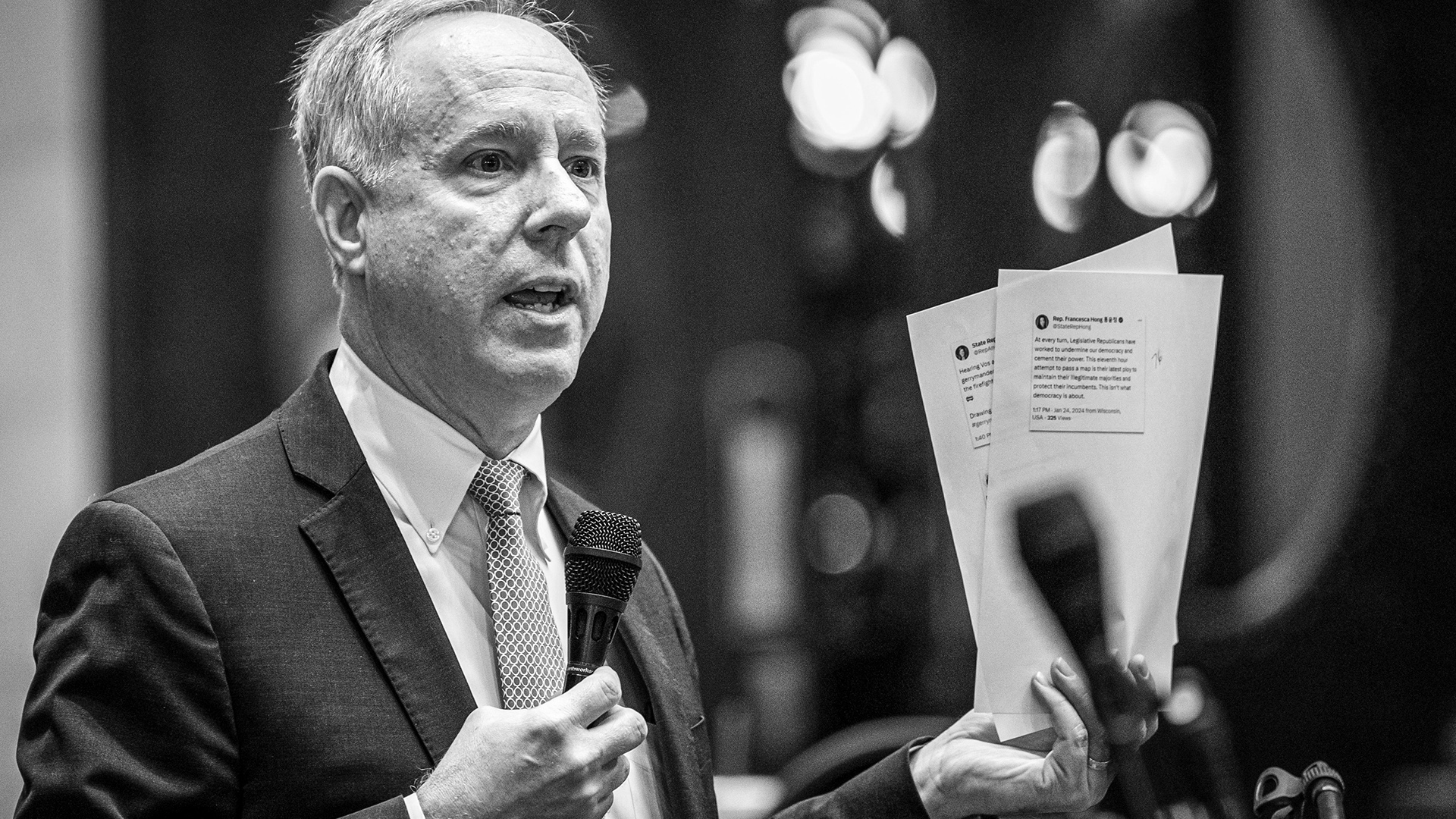 A grayscale photo shows Robin Vos standing and speaking while holding a microphone in his right hand and two sheets of paper in his left hand.