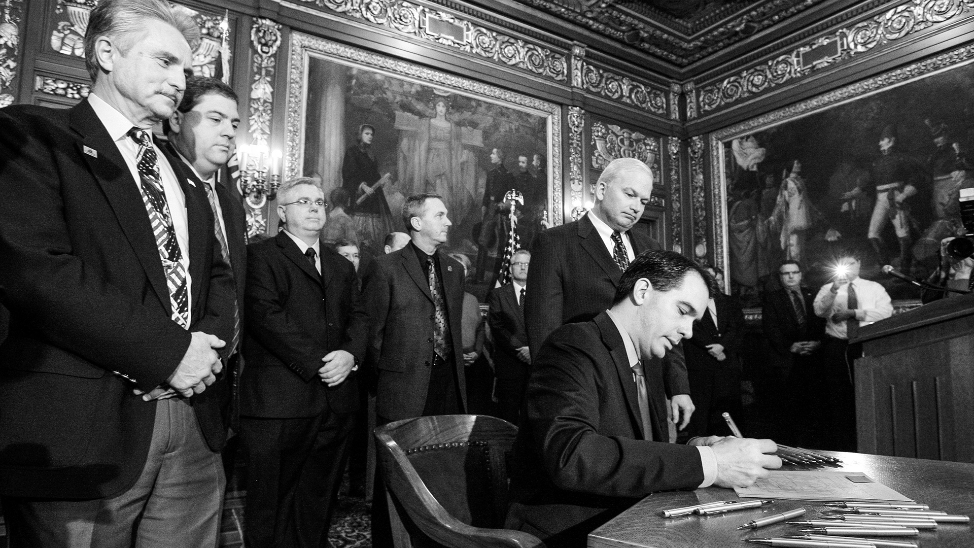 Scott Walker sits in a wood curved arm chair and holds a pen while signing a paper on a wood table, with multiple other pens on its surface, with Scott Fitzgerald and other people standing behind him, in a room with paintings and gilt filigree decorations on the walls and ceiling.