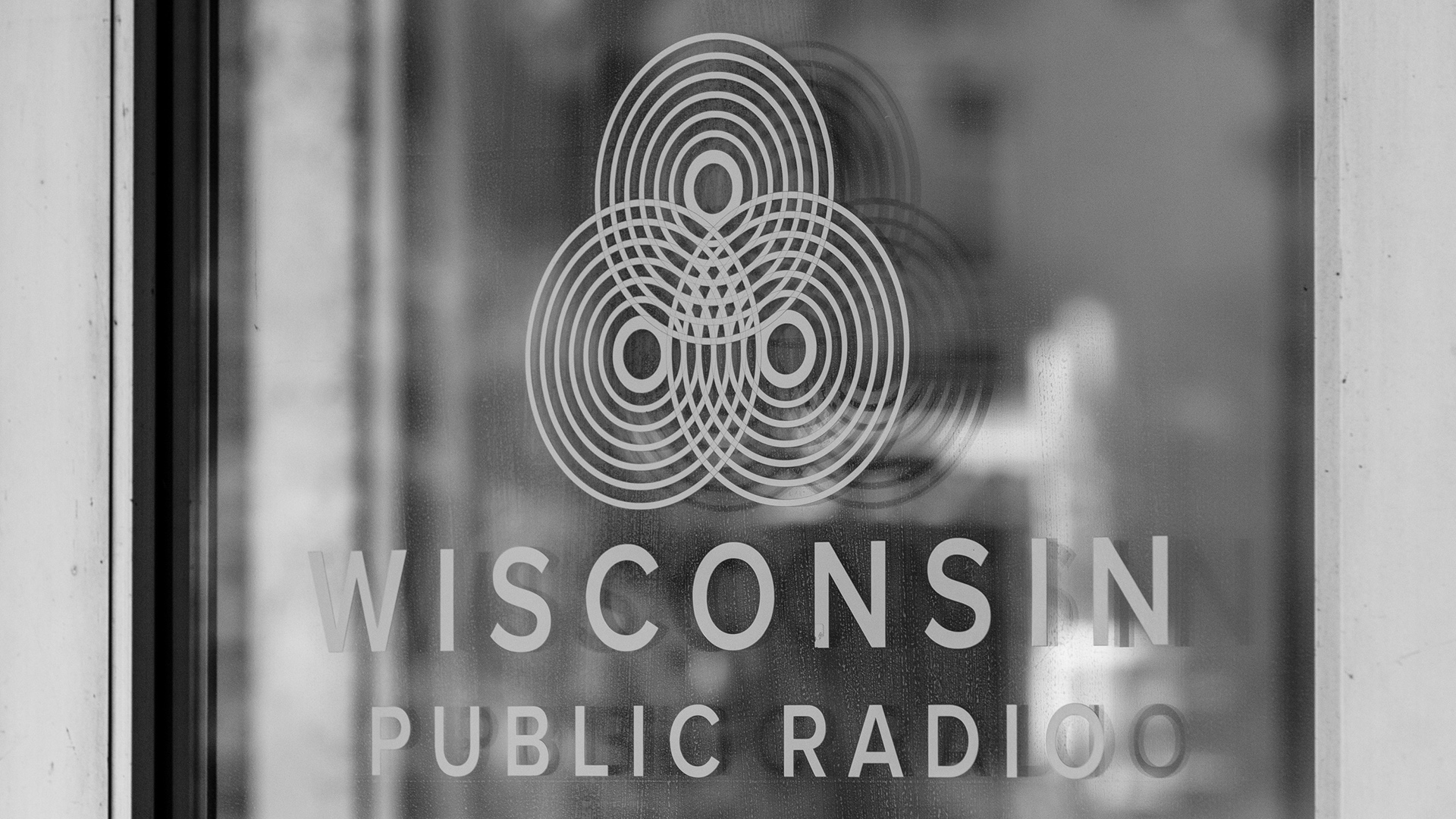 A grayscale photo shows a decal of the logo of Wisconsin Public Radio on a glass door.