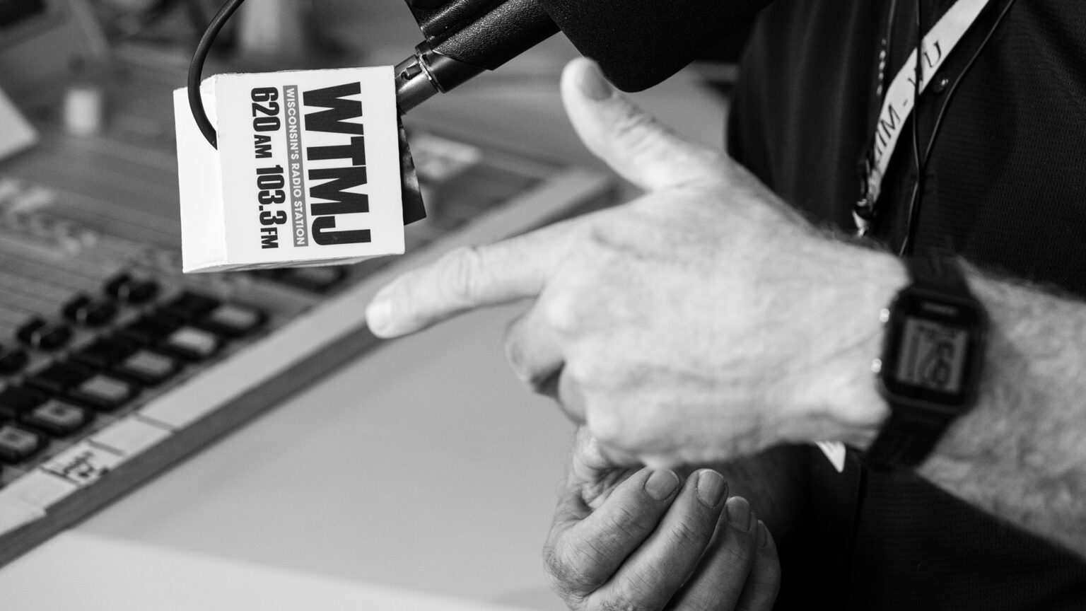 Two hands gesture beneath a microphone with a flag with the words WTMJ, Wisconsin's Radio Station and 620 AM 103.3 FM, with an out-of-focus broadcast radio console on the surface of a table in the background.