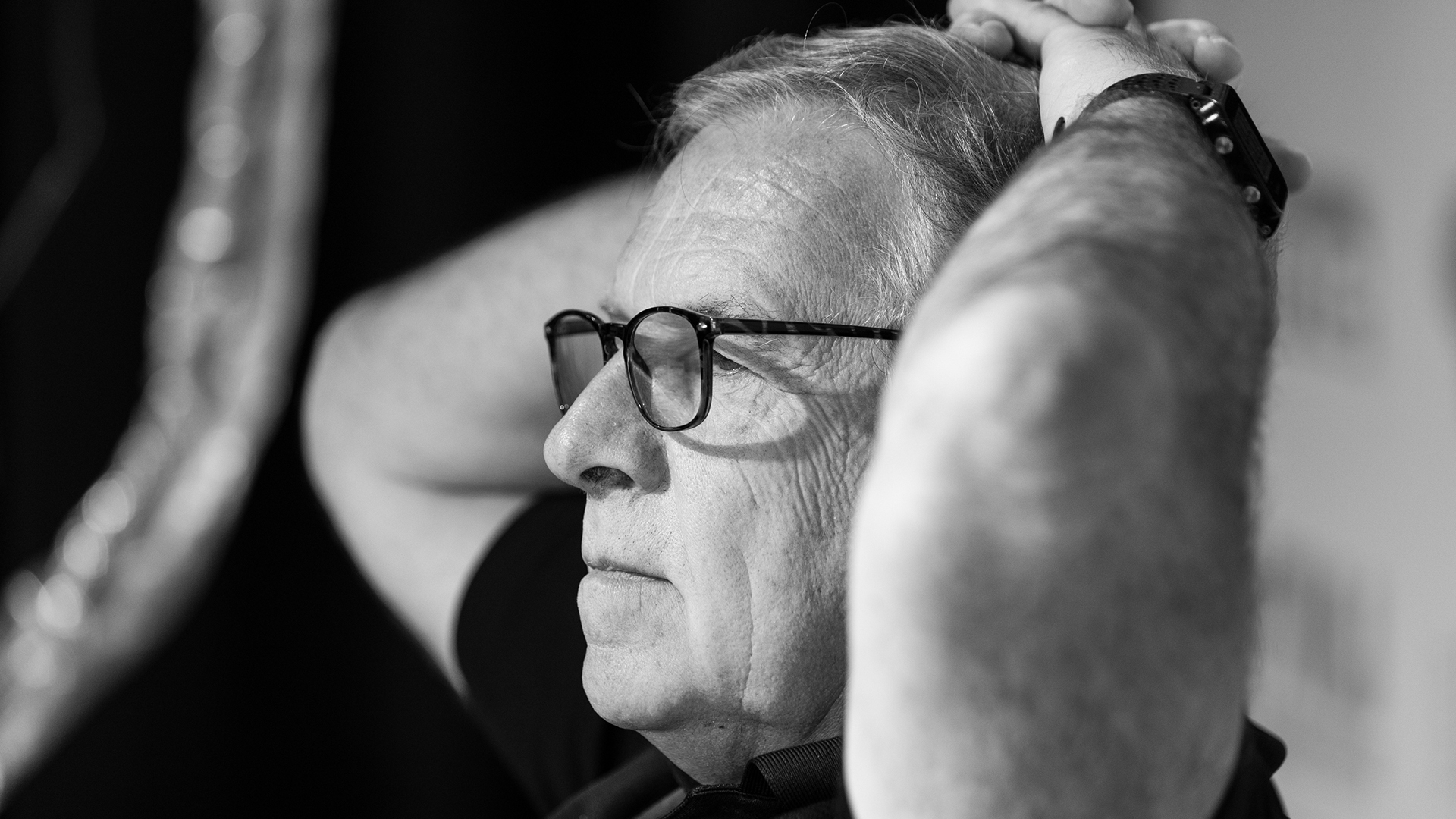 Steve Scaffidi sits with his fingers crossed atop his head, with an out-of-focus object in the foreground.