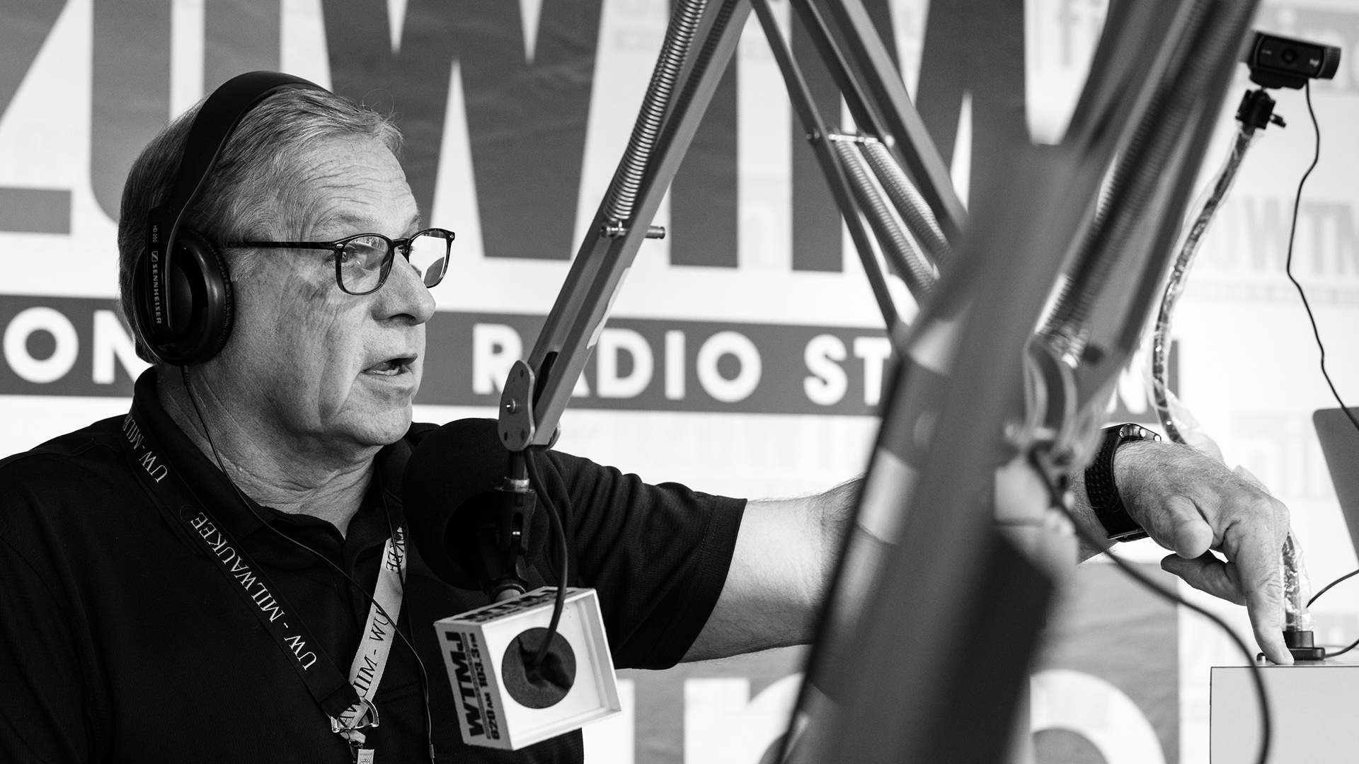 Steve Scaffidi pushes an audio operation button with his left hand while speaking into a mounted microphone with a WTMJ flag, with an out-of-focus microphone arm in the foreground.