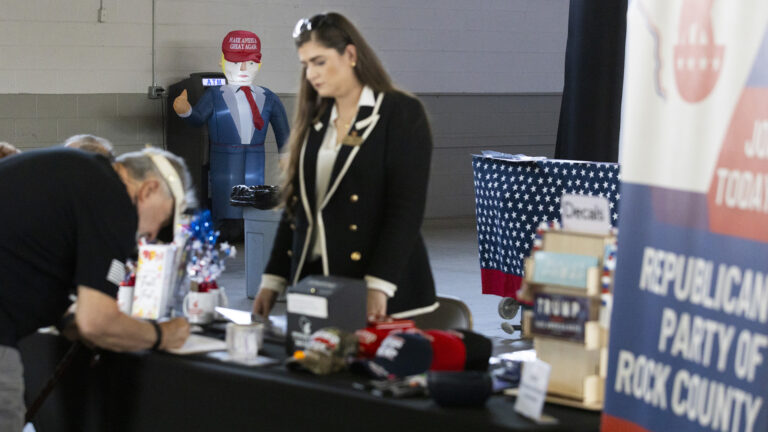 An inflatable doll of a figure costumed as Donald Trump with a Make America Great Again baseball cap stands in front of an ATM machine standing in front of a painted concrete wall, with out-of-focus people standing on opposite sides of a table with multiple items on its top in the foreground alongside an out-of-focus vinyl banner that includes the words Republican Party of Rock County.