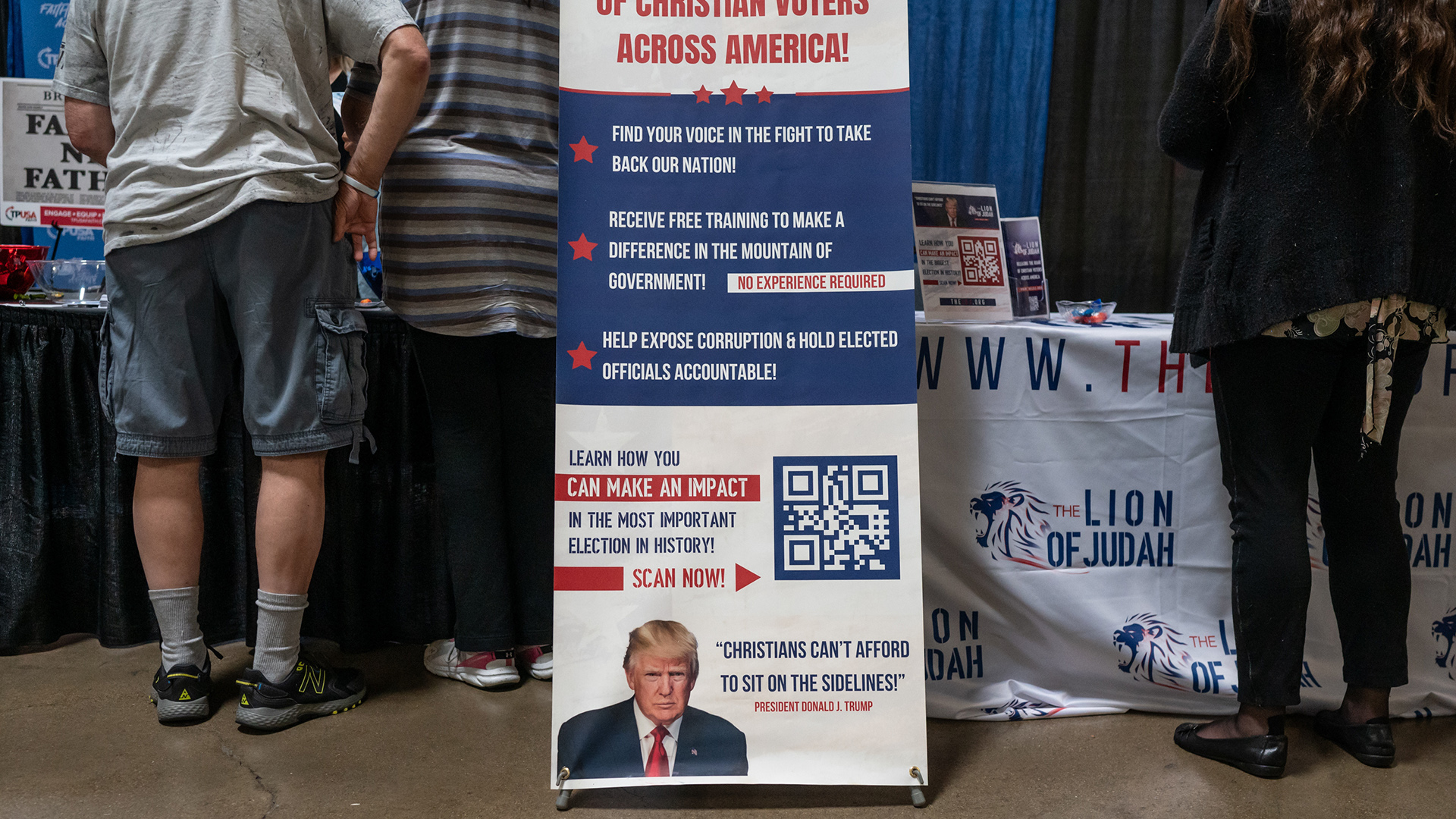 A vinyl banner attached with zip ties to a mount stands on a poured concrete floor in front of two tables, one with a tablecloth featuring the logo of The Lion of Judah, with people standing and facing the tables.