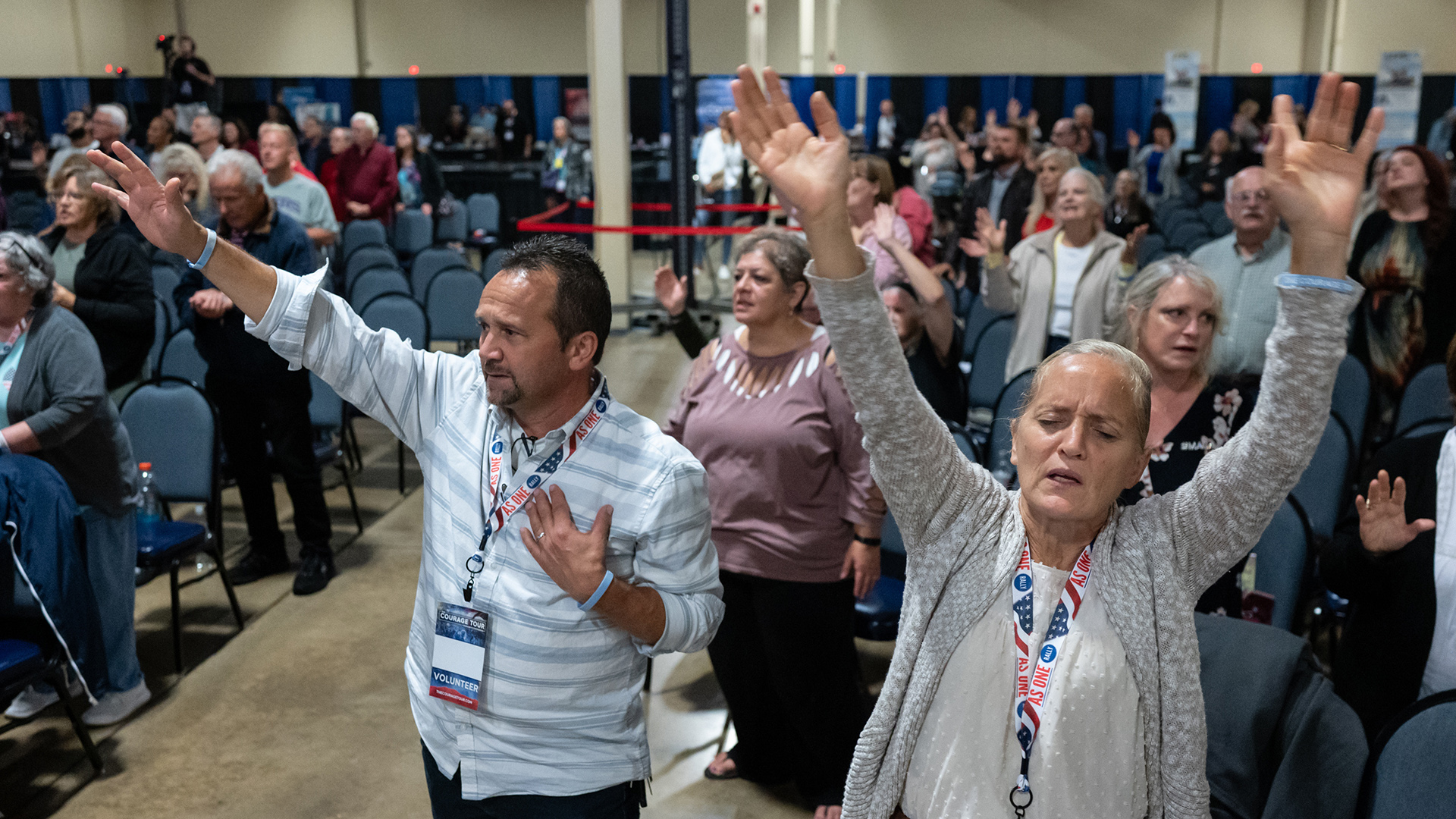 Manny King stands with his right hand raised and left hand over his heart and Mary Ann King stands with both arms raised in front of other people standing in front of rows of stackable chairs in a large room with support pillars and illuminated "EXIT" signs.