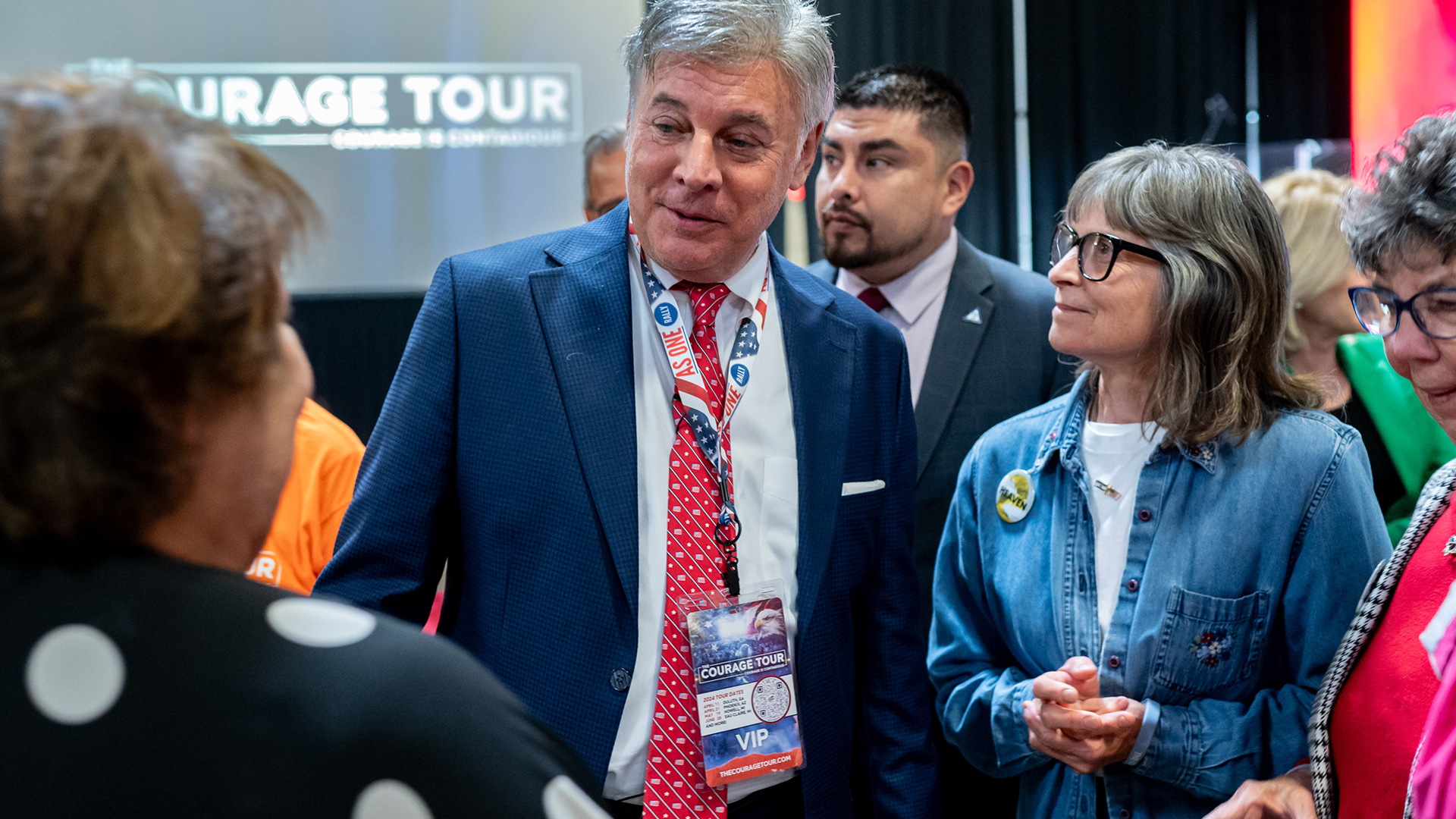 Lance Wallnau stands and talks to different people facing and standing next to him, in a room with a drop down projector screen in the background.