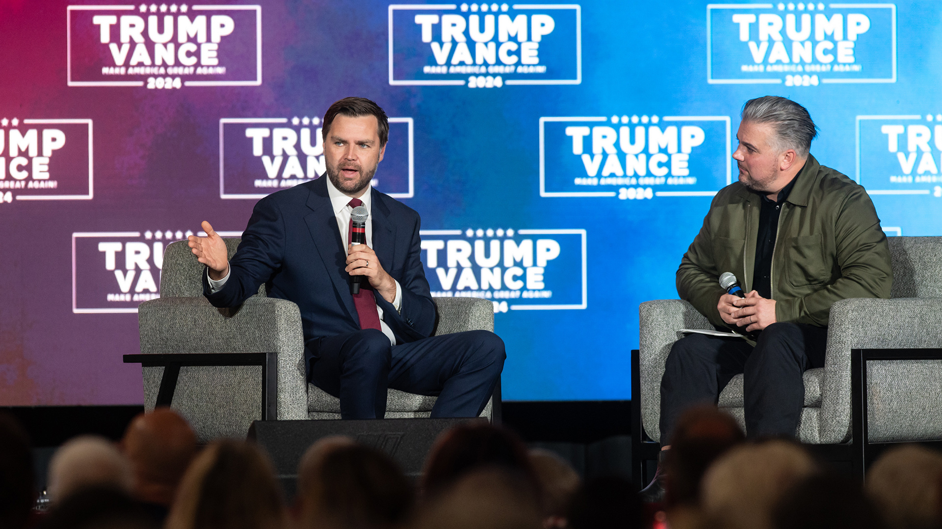 JD Vance sits in an upholstered armchair on a stage and speaks while holding a wireless microphone in his right hand and gesturing with his left hand, while another person seated in an adjacent chair watches, with a projector screen showing the Trump Vance 2024 campaign logo in the background and out-of-focus heads of audience members facing the stage in the foreground.