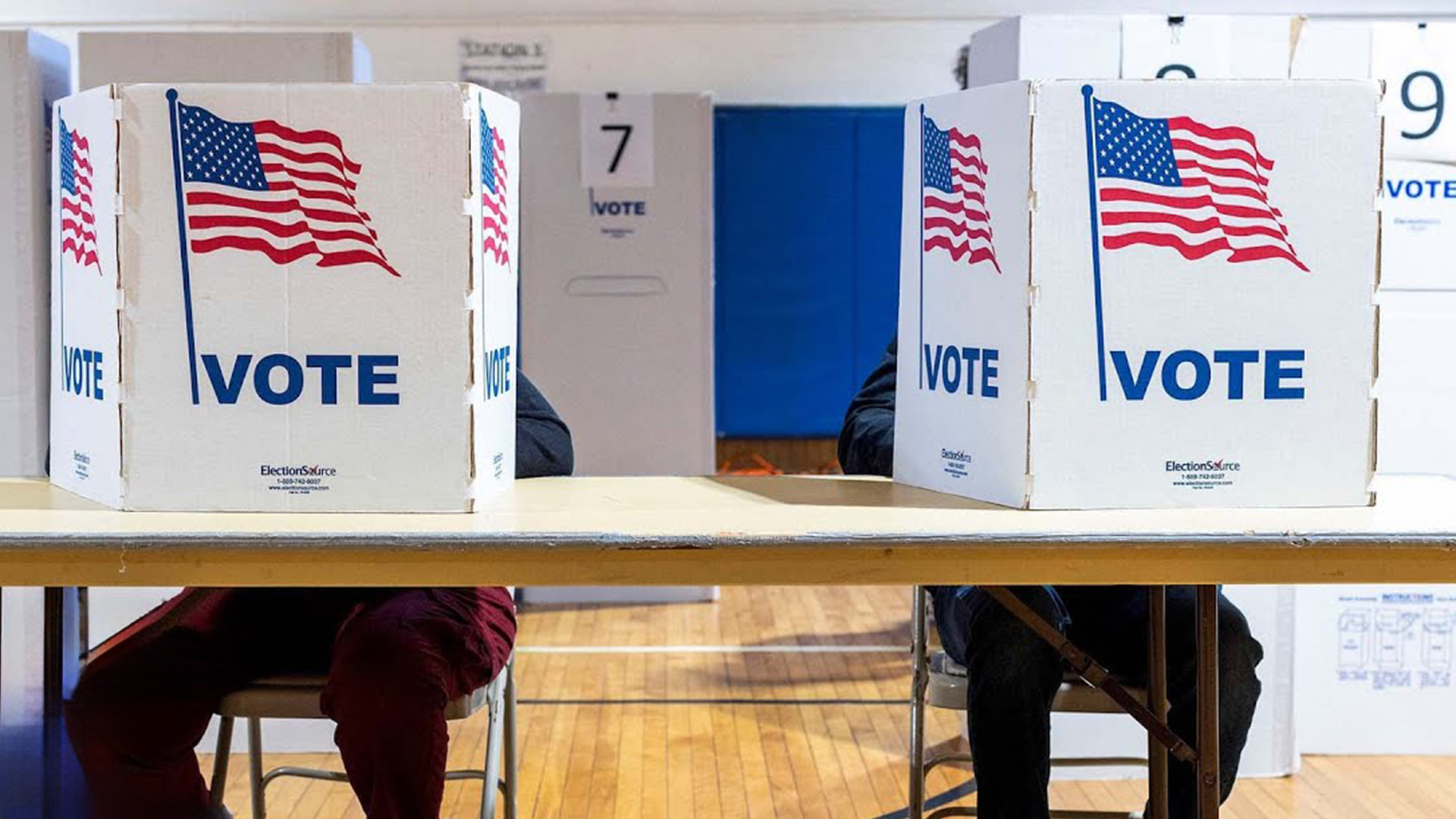 Two voters at a table in a polling place obscured behind cardboard partitions with graphic images of the American flag and the word "Vote" printed on them.