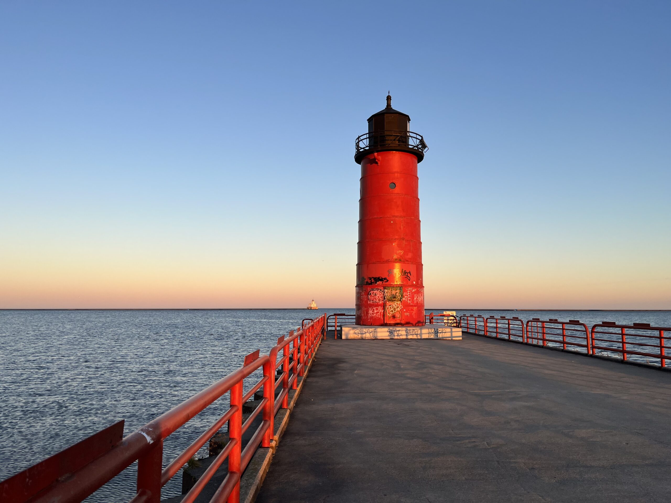 Milwaukee Pierhead Lighthouse at sunset with red fence running along pavement path connecting the lighthouse to the city.