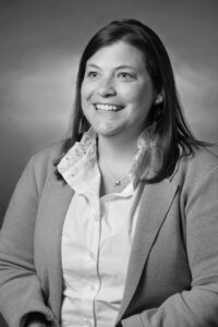 A grayscale photo featuring a portrait of a women from the waste up with shoulder length brown hair, wearing a blazer and white shirt with ruffled collar.