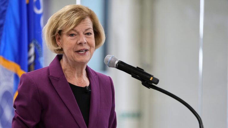 Tammy Baldwin speaks into a mounted microphone while standing in a room with a Wisconsin flag in the background.