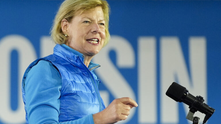 Tammy Baldwin gestures with her right hand while standing and speaking into a mounted microphone, with an out-of-focus sign in the background.