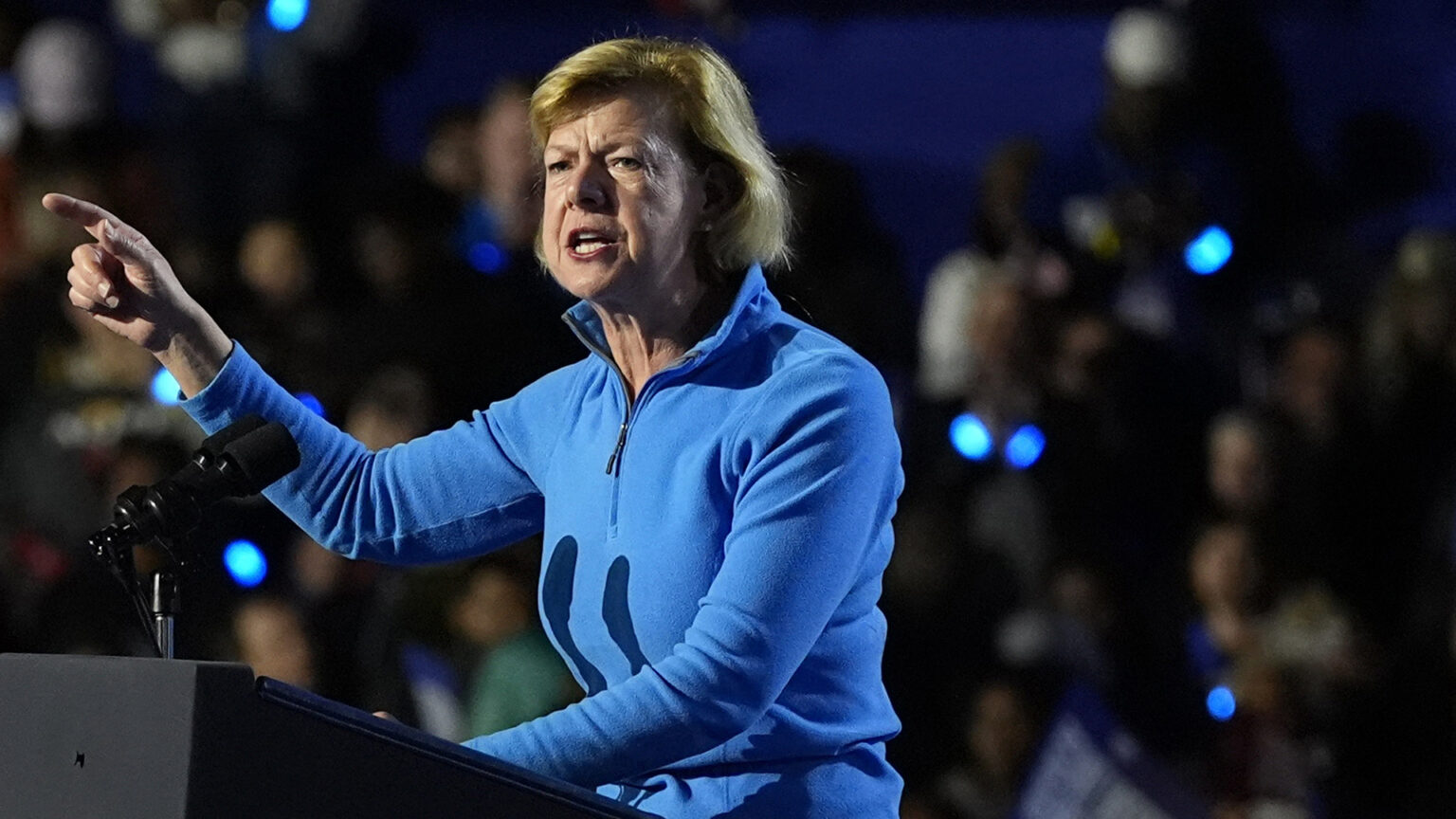 Tammy Baldwin gestures with her right hand while standing at a podium and speaking into a microphone mounted to its top, with out-of-focus people in the background.