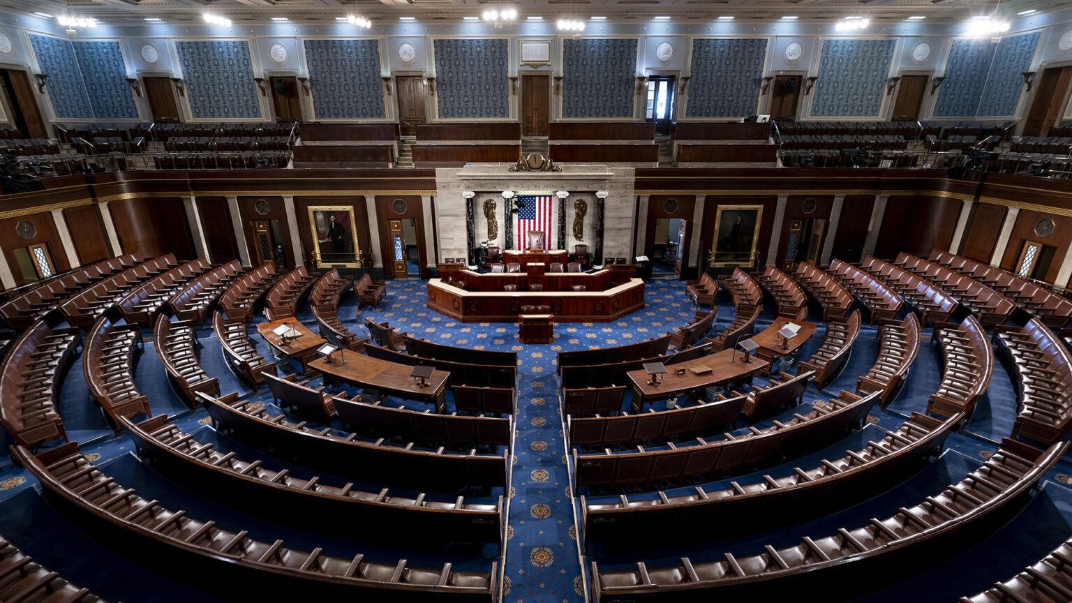 Multiple curved rows of connected chairs are arranged in a semicircle around a legislative dais with a U.S. flag on a wall behind it, with several tiers of seats ringing the chamber in an upper-level balcony.