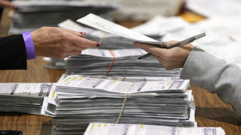 The hand of one person, holding a pen, hands a ballot to the hand of another person, over a wood table with multiple stacks of absentee ballot envelopes on its surface.