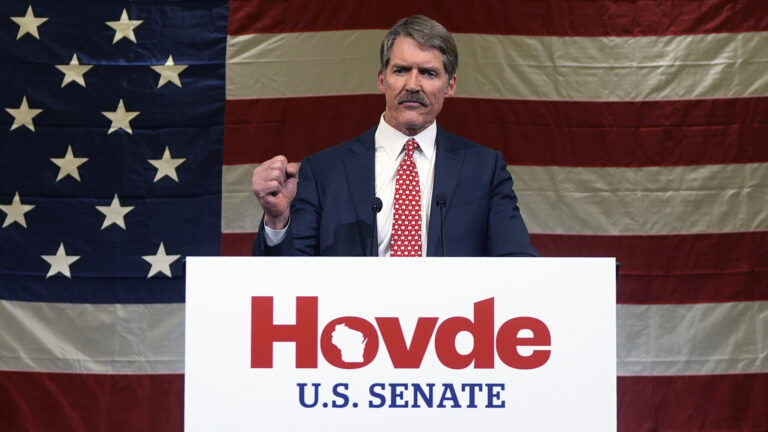 Eric Hovde makes a fist with his right hand while speaking into two microphones mounted to the top of a podium with a sign on the front with the words Hovde and U.S. Senate, with a large U.S. flag behind him.