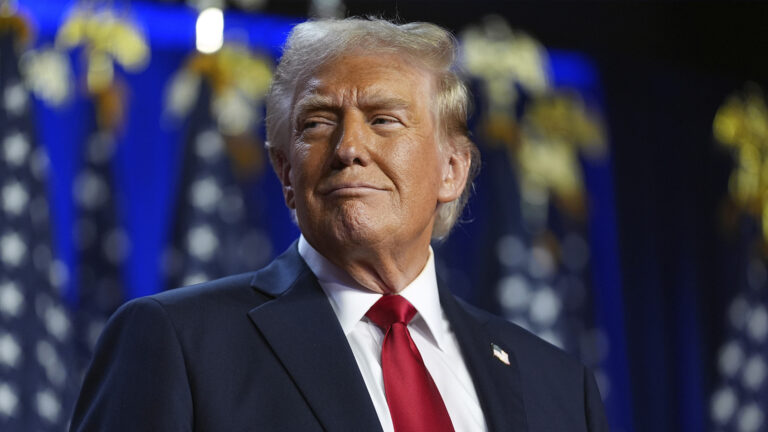 Donald Trump smiles while standing in a room with an out-of-focus row of U.S. flags in the background.