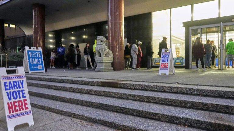 Multiple sandwich board sign with the words Early Vote Here and Vote Early stands at the bottom an top of a short set of marble steps , with people standing in a line next to two marble pillars and a stone statue of an animal next two a building with a glass wall and double doors open to an illuminated interior.