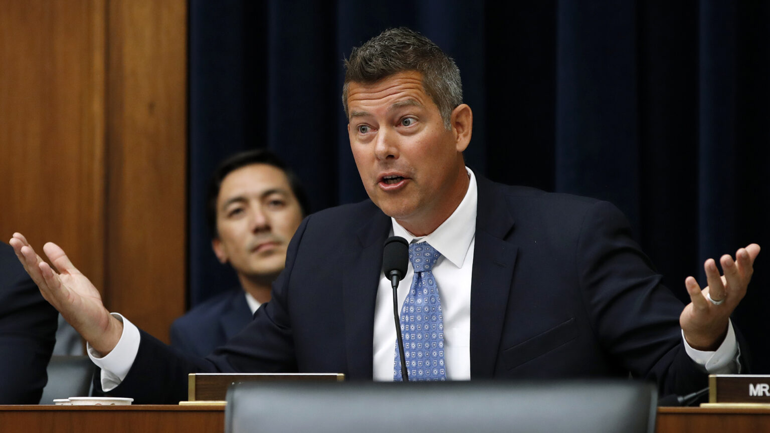 Sean Duffy gestures with both hands while seated and speaking into a microphone mounted to a wood desk, with an out-of-focus person seated in the background.