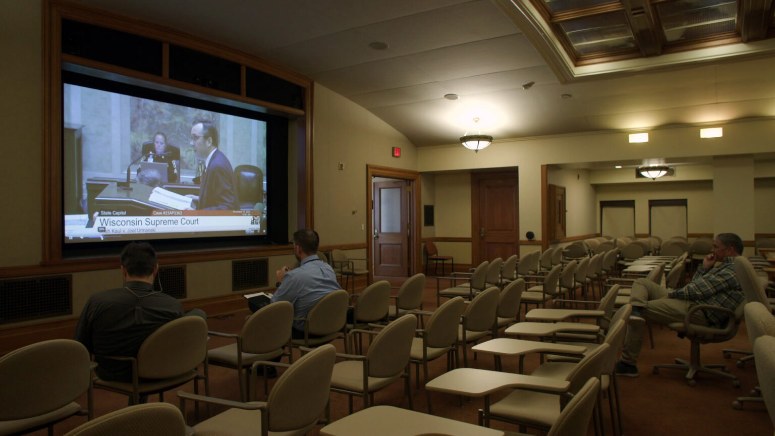 Three people seated in rows of stackable chairs, desk chairs and a swiveling office chair sitting in a room with a skylight and illuminated light fixtures watch a projection screen bordered by wood moulding shows a video feed of a man standing at a podium with an open binder filled with papers on its surface, with another person seated in the background in a high-backed chair, with a graphic at the bottom with the text Wisconsin Supreme Court.