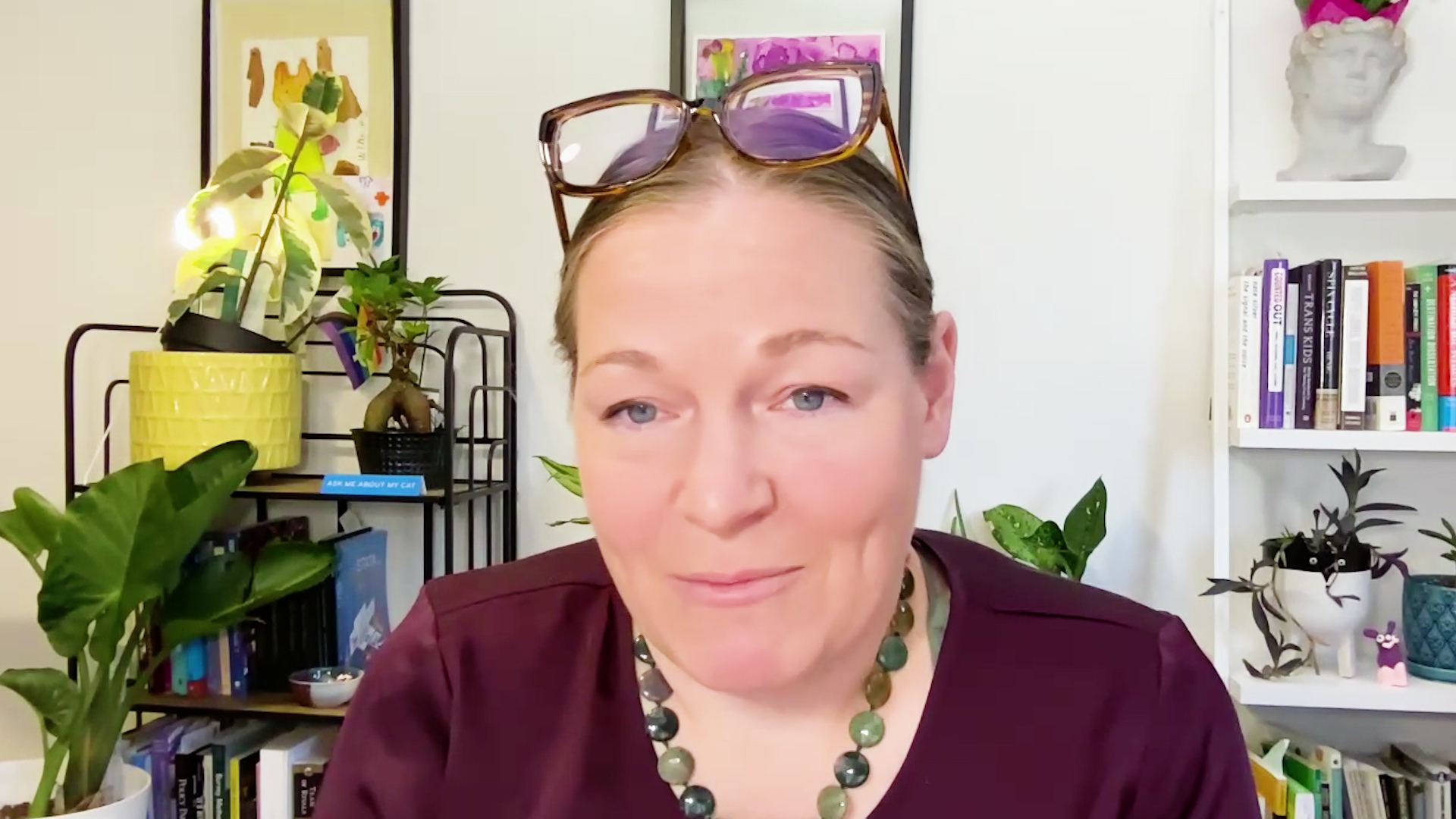 Mollie McQuillan sits in a room with a bookshelf lined with books, potted plants and other items, a cart with more books and potted plants, and framed artwork on the wall in the background.