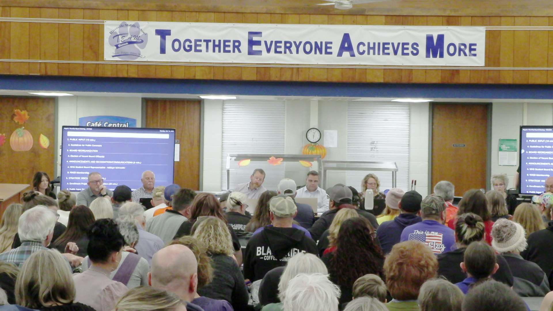 School board members sit at a table with numerous people seated facing them in a high ceiling room with wood paneled walls, TV monitors on either side of the room and a banner above reading "Together Everyone Achieves More," with the letters TEAM capitalized.