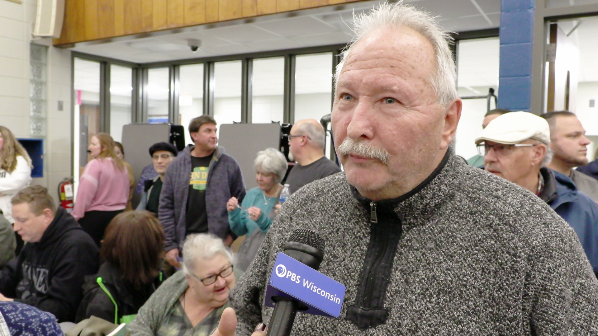 Gene Schmidt speaks into a microphone with a "PBS Wisconsin" flag while standing in front of multiple people standing and sitting with a wall of windows in the background.