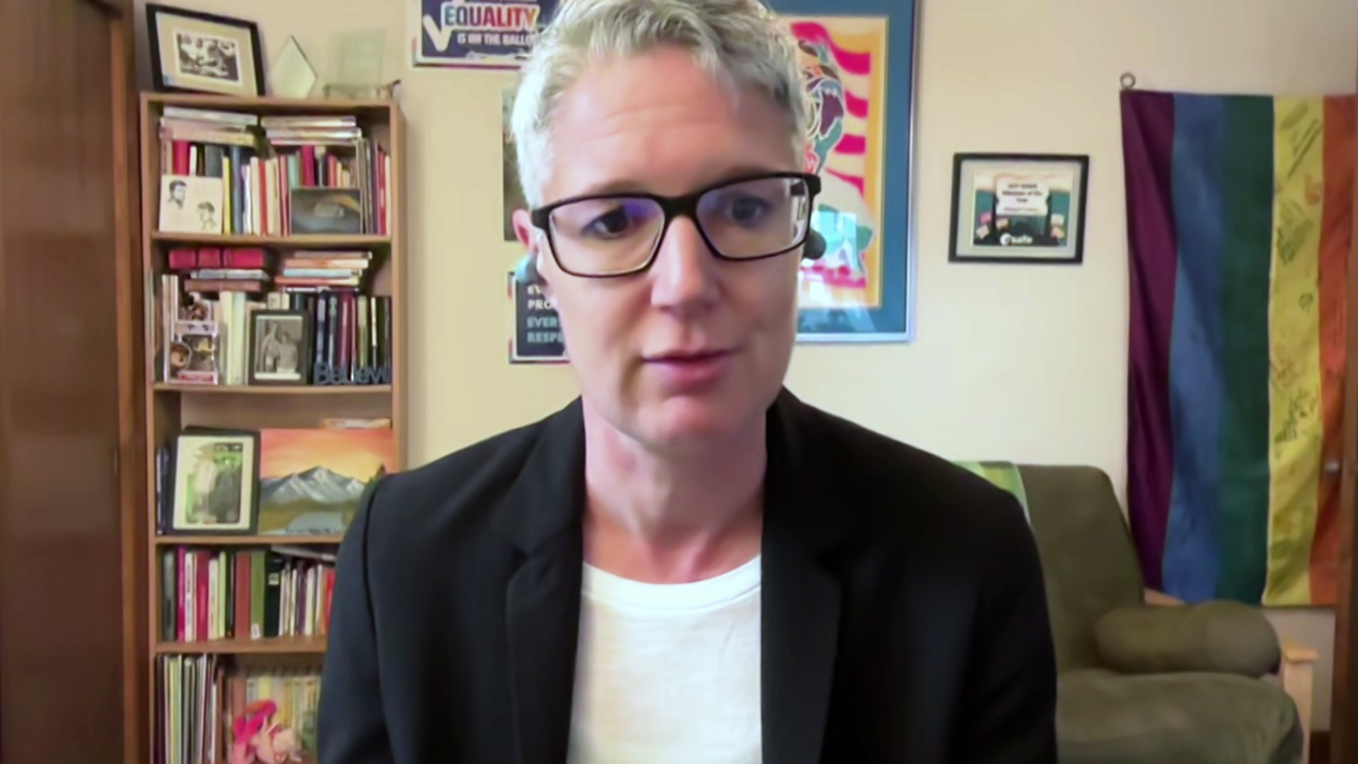 A still image from a video shows Abigail Swetz seated in a room with a bookshelf lined with books and other items, framed artwork on the wall, a rainbow Pride flag and an armchair in the background.