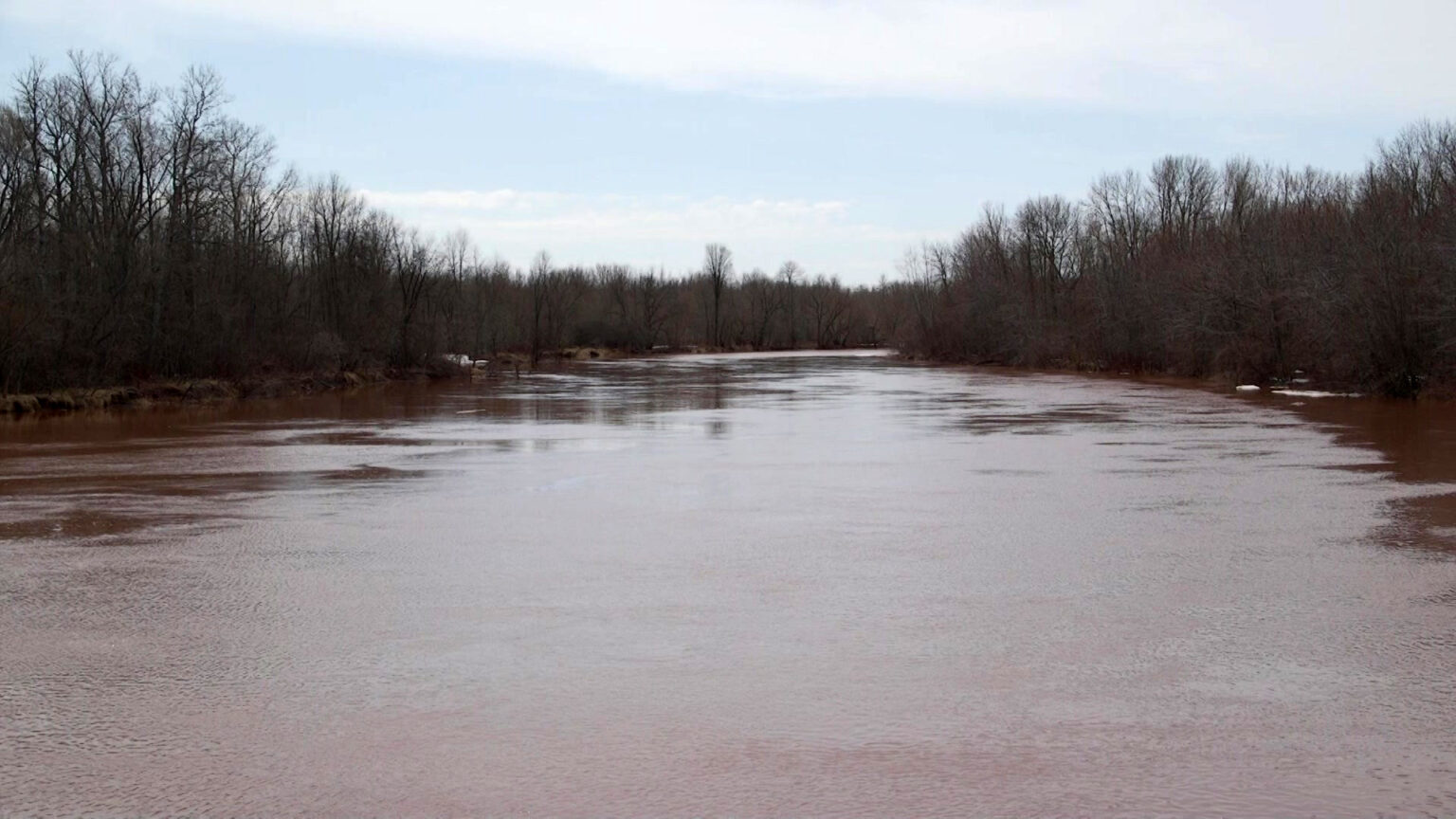 A river tinted red with iron-rich soil flows toward the horizon, with leafless trees on either side under a partly cloudy sky.