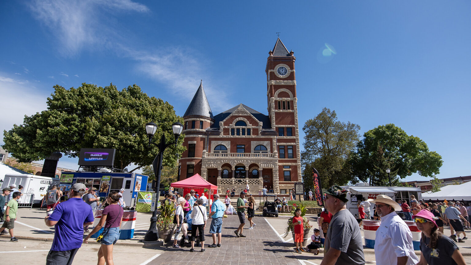 Pedestrians walk on sidewalks and streets among tents and food trucks in front of a brick-and-masonry Italianate  building with a conical tower and a clock tower, standing among multiple large deciduous trees.