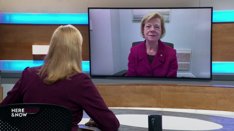 Frederica Freyberg sits at a desk on the Here & Now set and faces a video monitor showing an image of Tammy Baldwin.