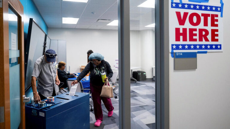 A voter at a polling place places an absentee ballot envelope into a drop box with multiple rolls of I Voted stickers on its surface and a poll worker standing behind it, with another voting standing at a table opposite a seated poll worker in a room with large monitors mounted to one wall, low-pile carpet and fluorescent panel lights in the ceiling, as seen through an open wood door with a glass window.