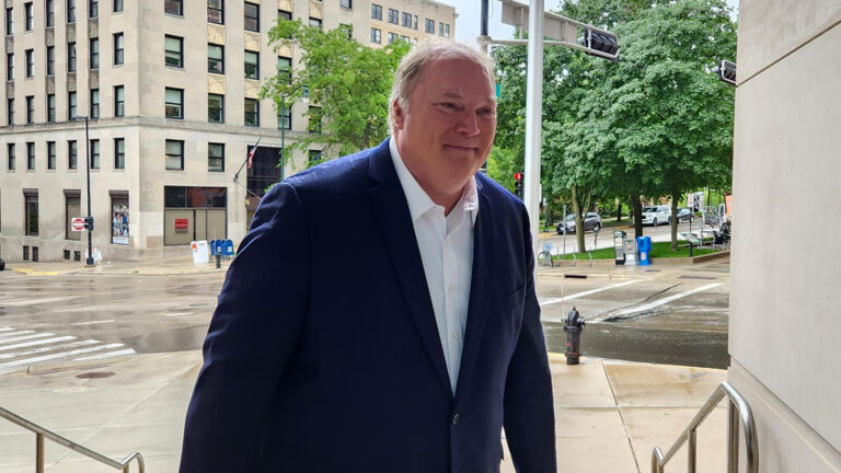 Michael Gableman walks up steps toward the entrance of a building from a sidewalk in front of an intersection, with buildings, trees, traffic signals and parked vehicles in the background.