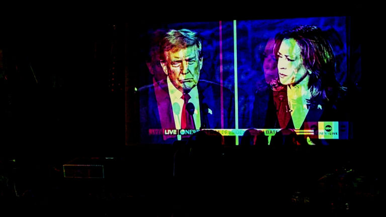 A photo illustration featuring 3-D style dual images in multiple colors shows the silhouettes of heads facing a projection screen showing a split-screen image of a debate between Donald Trump and Kamala Harris.