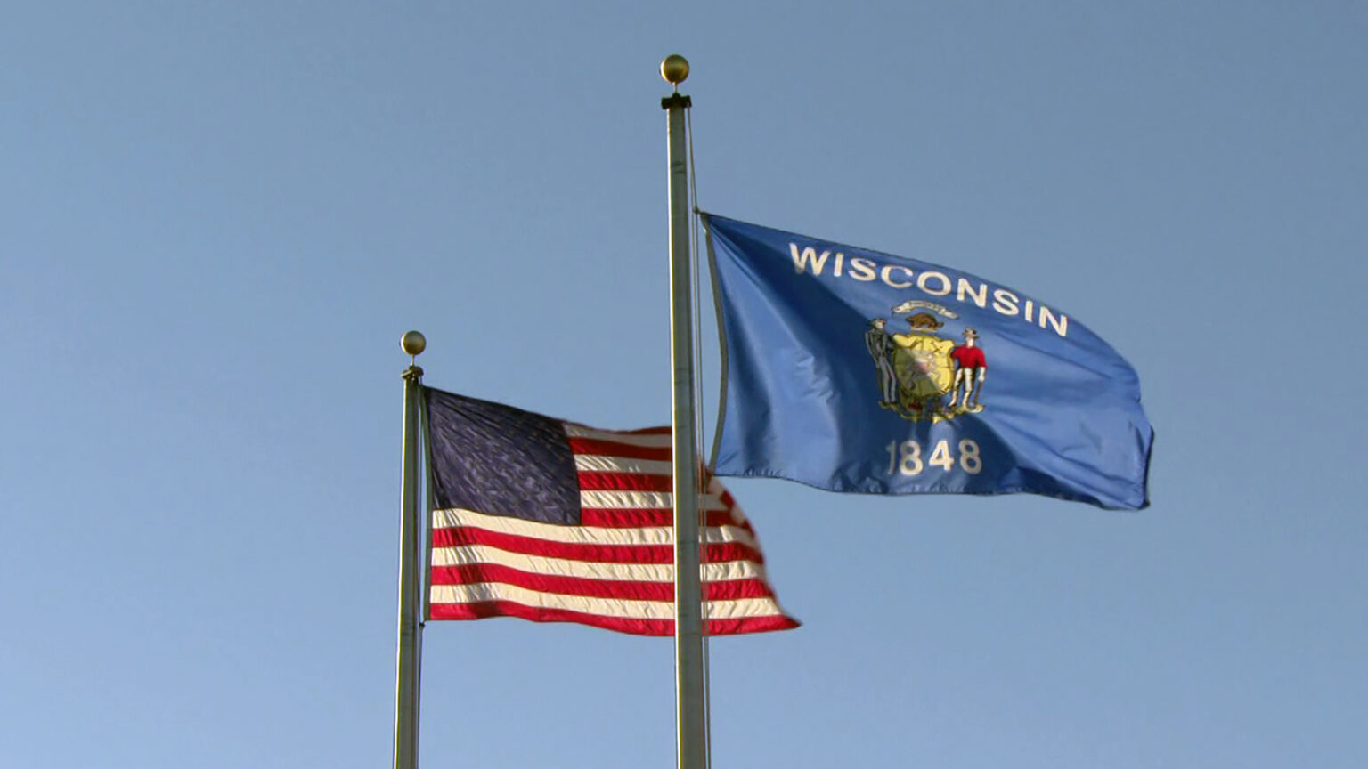 The U.S. and Wisconsin flags wave while attached to flagpoles, with a clear sky in the background.