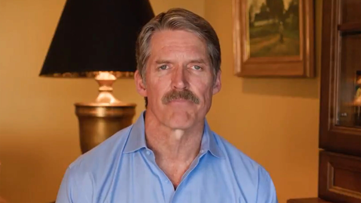 A screenshot of a video shows Eric Hovde sitting in a room with an illuminated lamp, wood-and-glass cabinet and wall-mounted painting in the background.