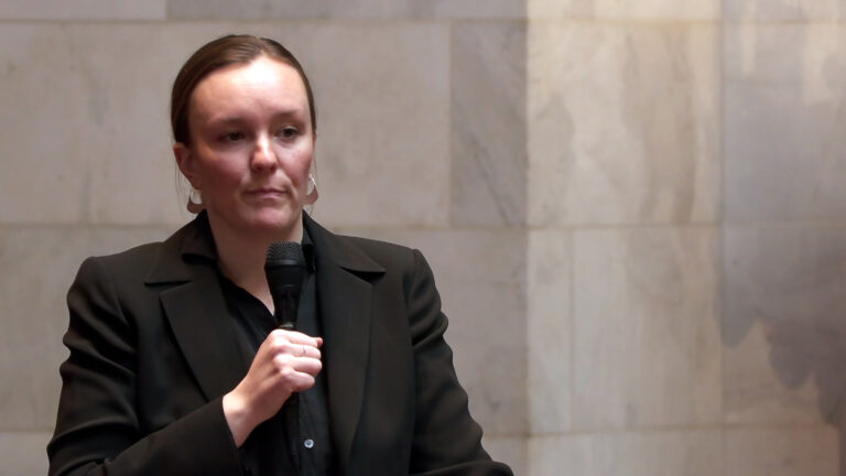 Greta Neubauer speaks into a wireless microphone while standing in a room with marble masonry in the background.