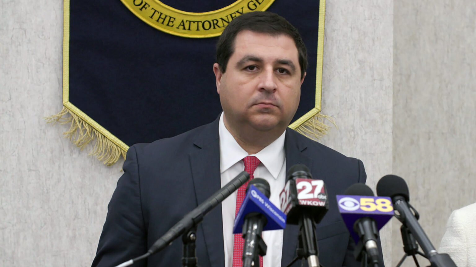 Josh Kaul stands and speaks into multiple microphones, several with the flags of different media organizations, in a room with a fringed velvet banner displayed on a wall in the background.