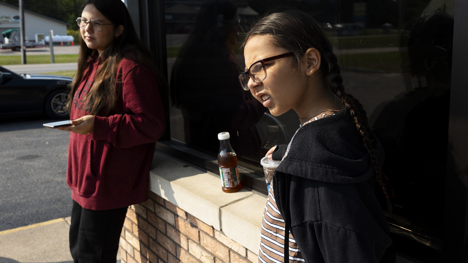 Sophie and Nate Hagen stand against a brick and concrete ledge outside a tinted window of a building, with a parked vehicle, road and building in the background on one side.