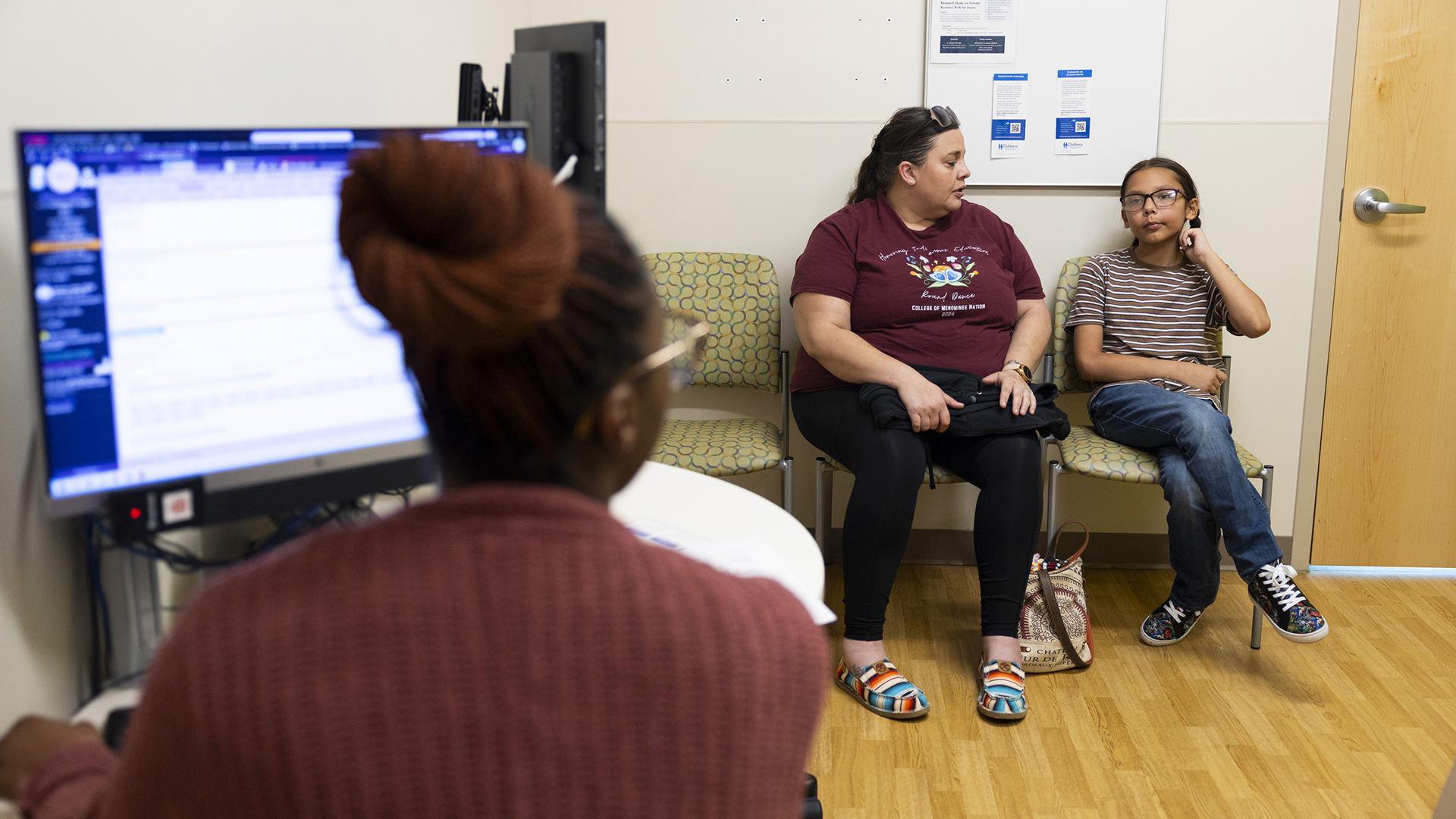 Lynda and Nate Hagen sit in stackable chairs against a wall and next to a door with a lever handle in a room with wood flooring, with an out-of-focus nurse seated next to a computer monitor facing them.