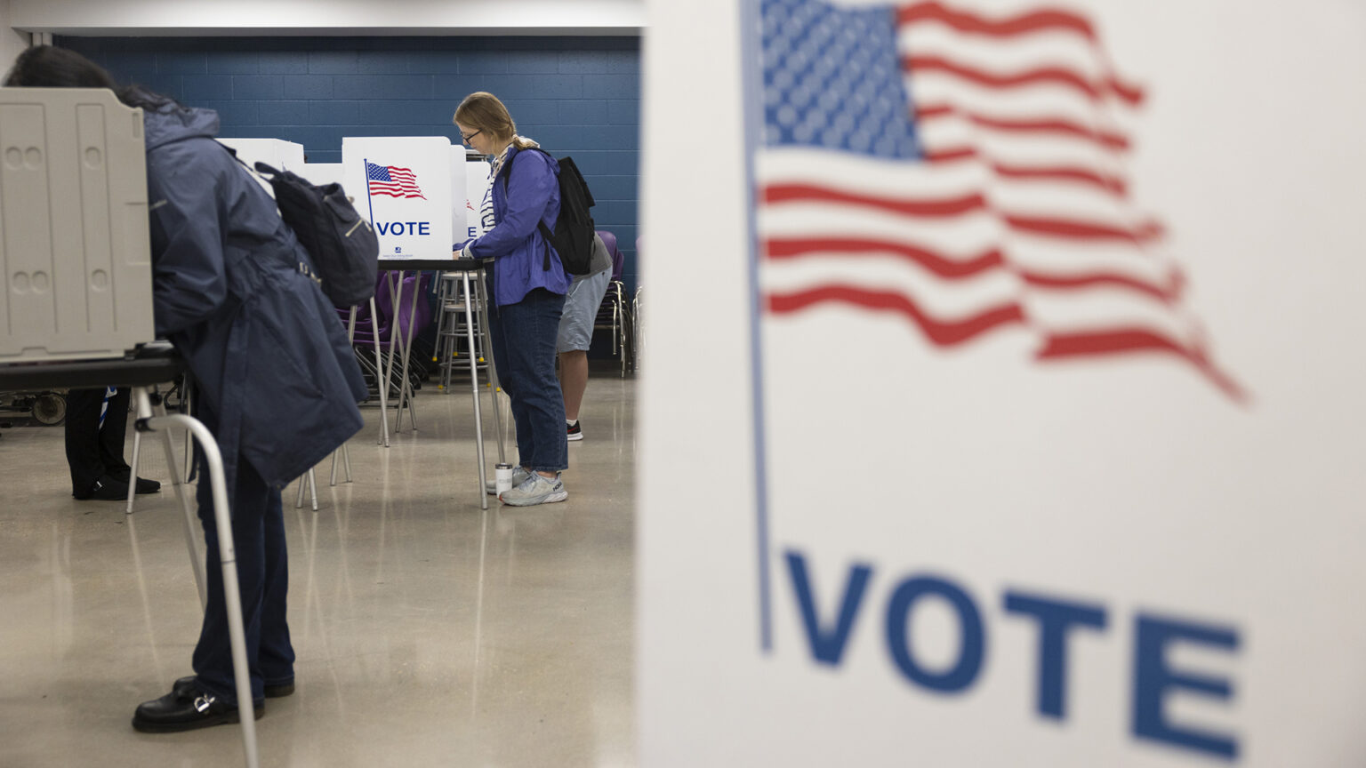 Voters stand and face temporary voting booths with privacy screens with a U.S. flag illustration and the word Vote on their side wall, a plastic surface and metal legs, in a room with painted concrete block walls and a linoleum floor, with an out-of-focus voting booth side wall in the foreground.