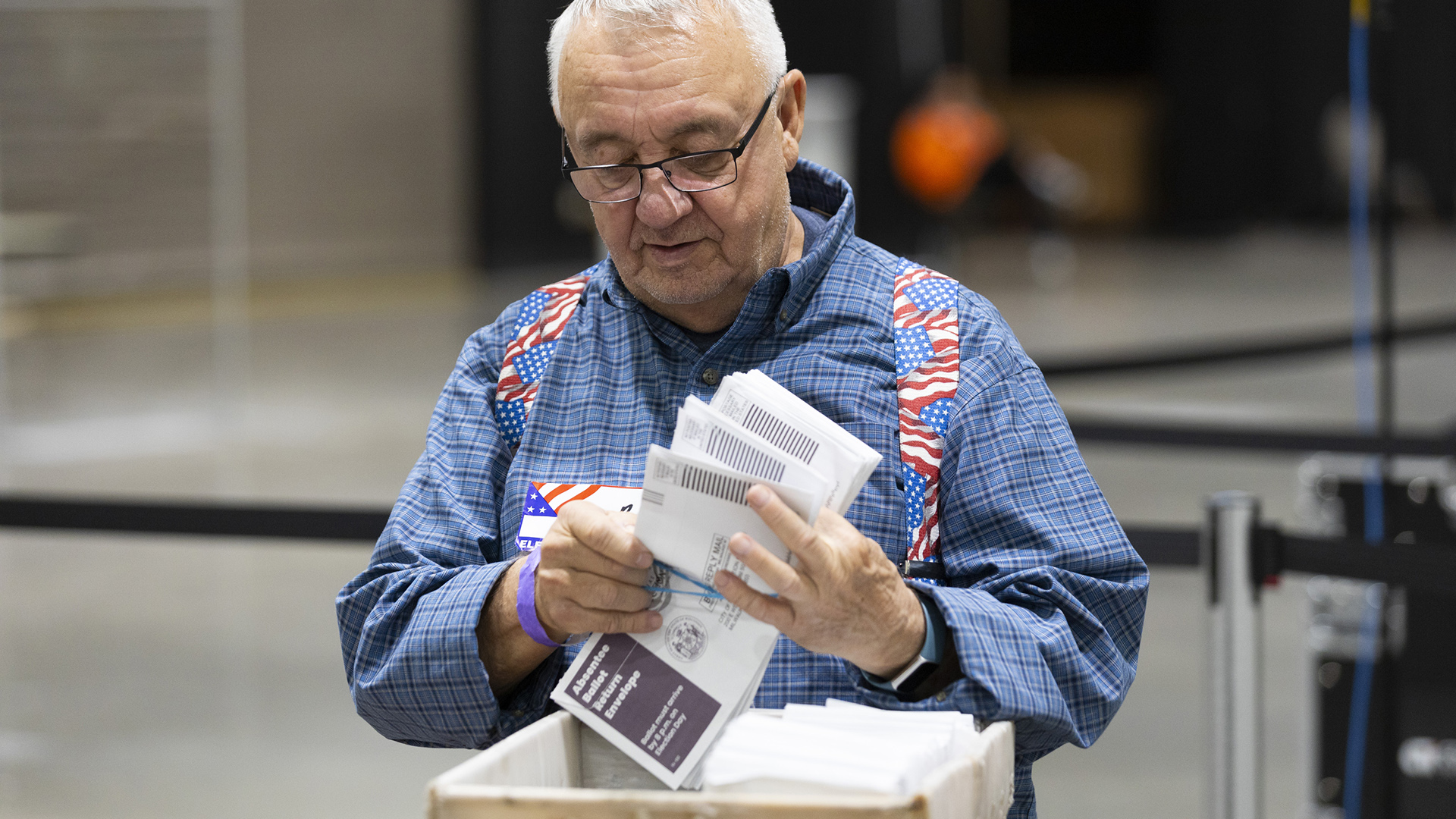 An election worker holds multiple absentee ballot envelopes in their hands above a box filled with more envelopes, with out-of-focus retractable belt barriers in the background.