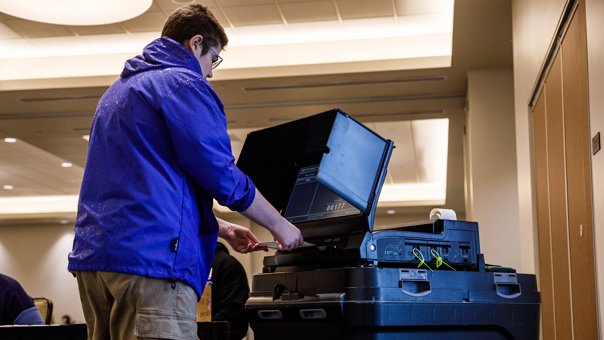 Theordorus Guigley submits a paper ballot into a ballot tabulation machine in a room with recessed fluorescent lighting in the ceiling.