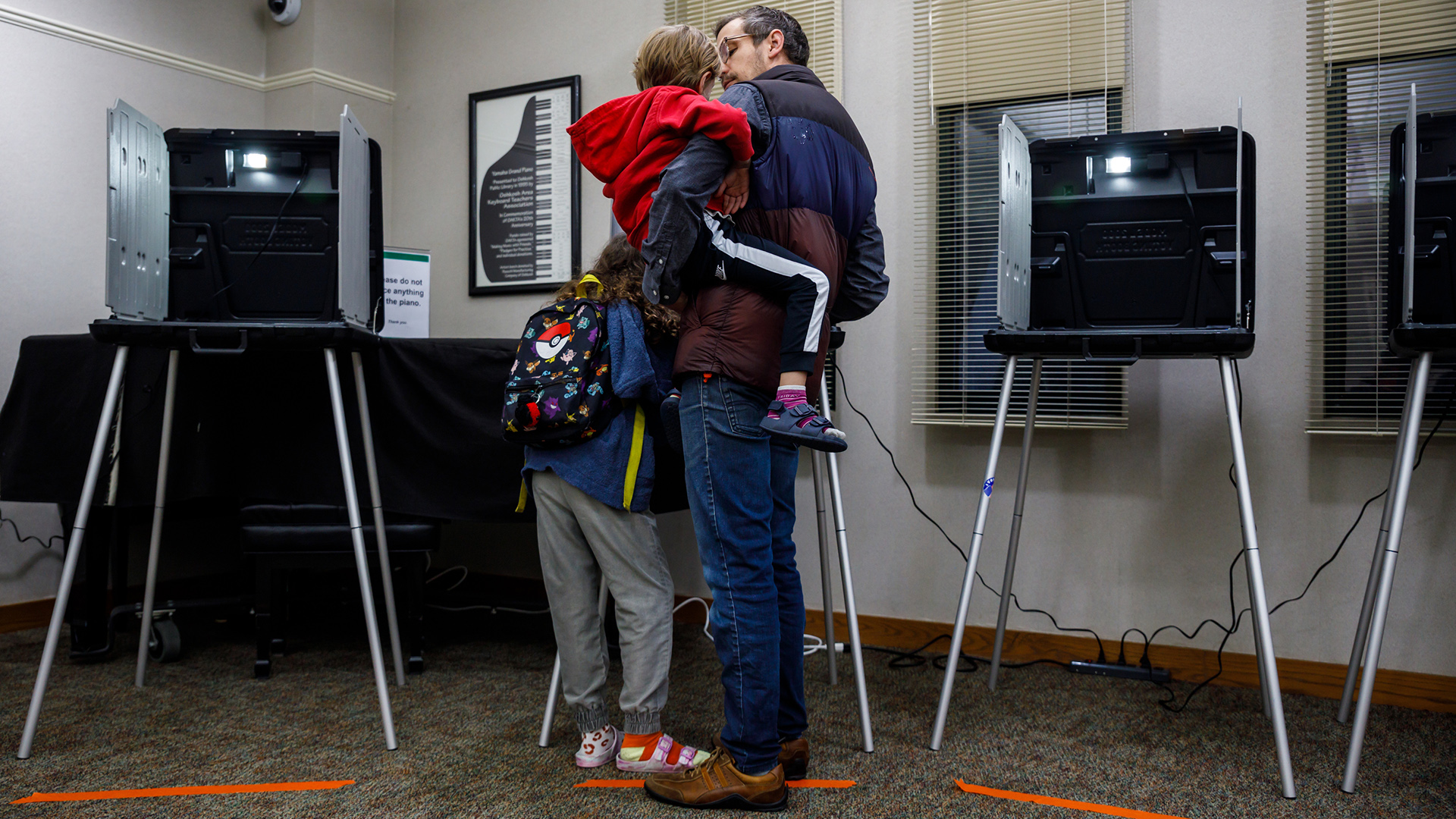 Dylan Chmura-Moore, holding one chlid in his arms and with another standing at his side, stands in front of a temporary voting booth with plastic privacy screens and table, and metal legs, with additional voting booths on either side and in front of a covered piano, in a room with a low-pile carpet and windows with lowered open blinds.