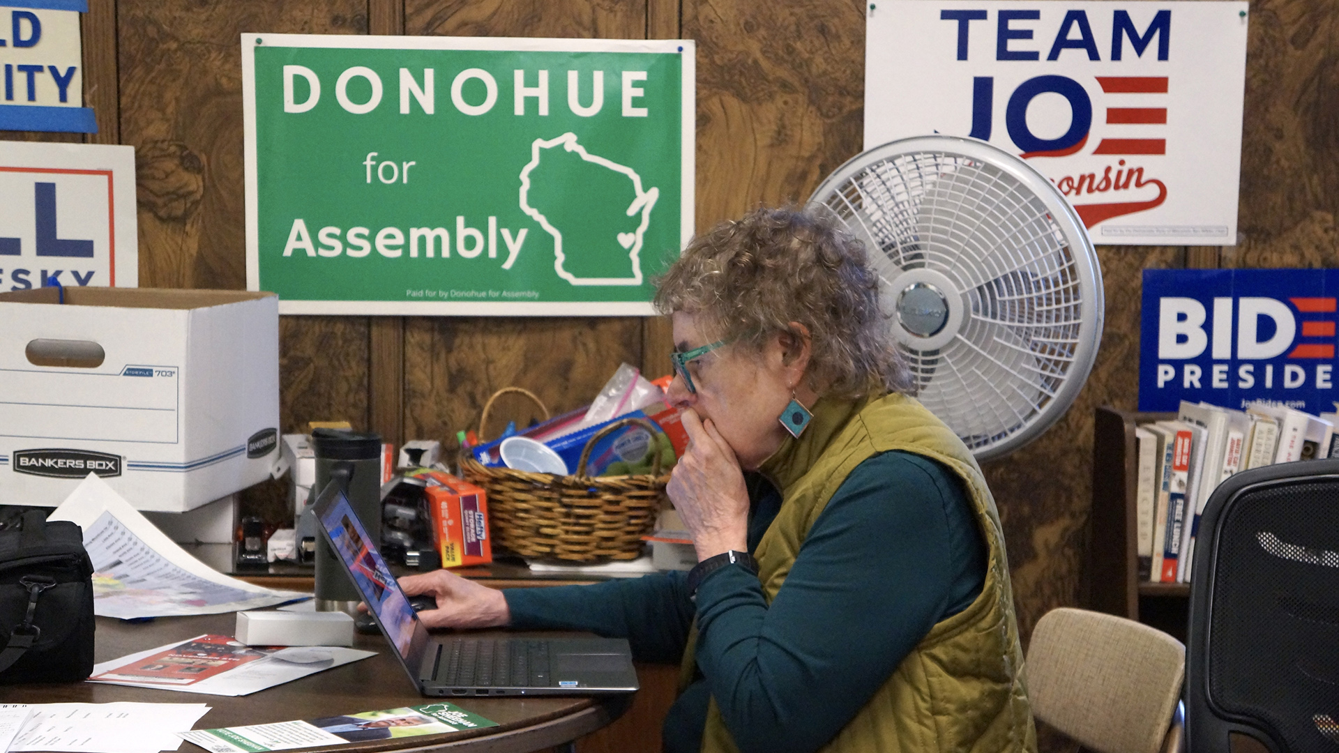 Mary Lynne Donohue sits on a folding chair and looks at a laptop computer on a round table, with political flyers, office supplies, a banker's box and other items on its surface, with a low bookshelf and multiple political yard signs pinned to a wall in the background.